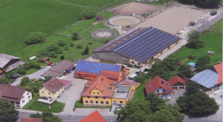 Luftaufnahme einer ländlichen Gegend mit Solarmodulen auf den Dächern, von Grün umgebenen Gebäuden und einer großen Arena, die eine idyllische Startseite-Szene zeigt.