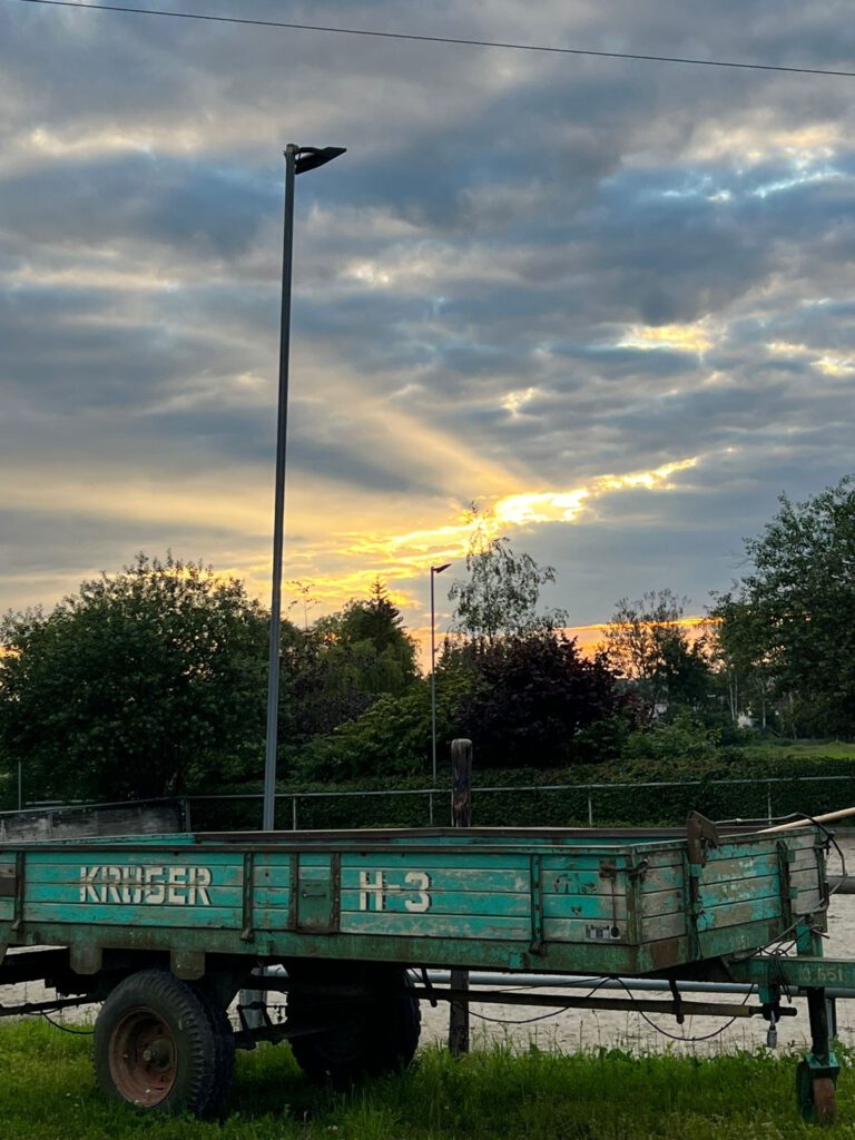 Rustikaler blaugrüner Wohnwagen auf einem Feld bei Sonnenuntergang mit dramatischen Wolken und einer einzelnen Straßenlaterne, die Fernweh weckt. Bäume im Hintergrund vervollständigen diese ruhige Startseite.