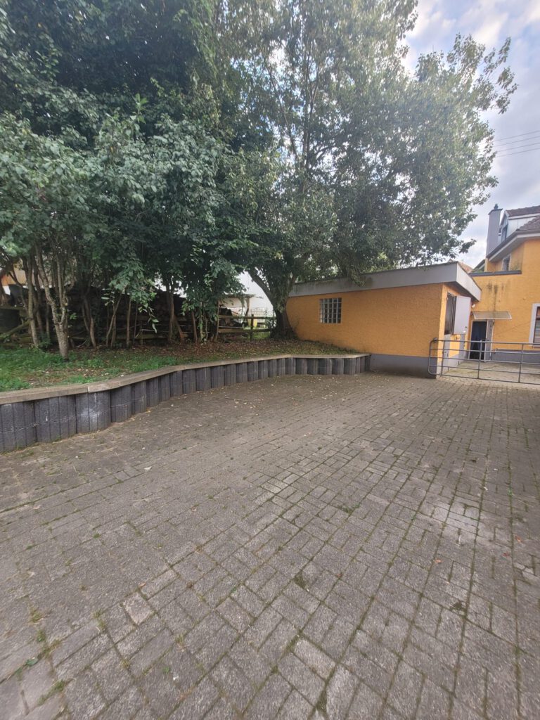 Gepflasterter Innenhof mit erhöhtem Sitzbereich aus Stein, gesäumt von Bäumen und angrenzend an gelbe Gebäude, die eine charmante Startseite zum Entspannen bilden.