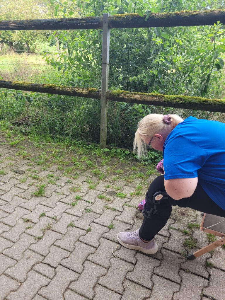 Eine Person mit einer Knieorthese kniet auf einem gepflasterten Weg in der Nähe eines Gartens und bindet die Schnürsenkel eines Kindes.