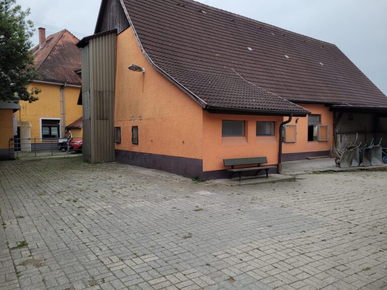 Ein großes orangefarbenes Gebäude mit Ziegeldach und einer Bank davor, gelegen in einem Innenhof mit einigen anderen Gebäuden in der Nähe, dient als perfekte Startseite für Besucher.