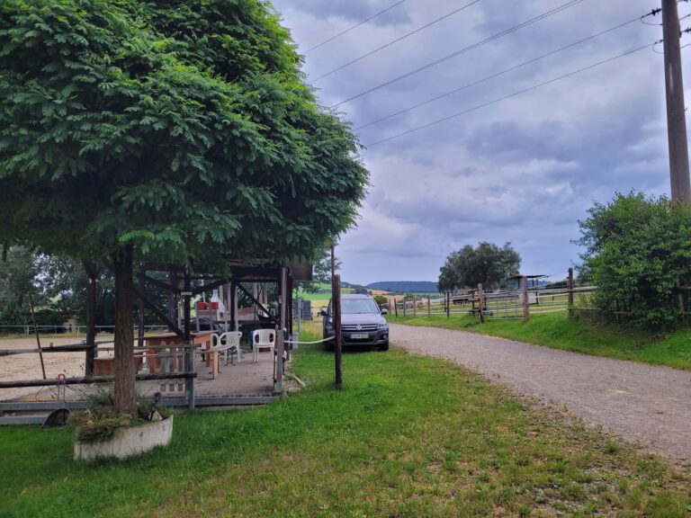 Eine ländliche Startseite mit einer baumbestandenen Terrasse, einem geparkten Auto, einem Kiesweg und bewölktem Himmel.