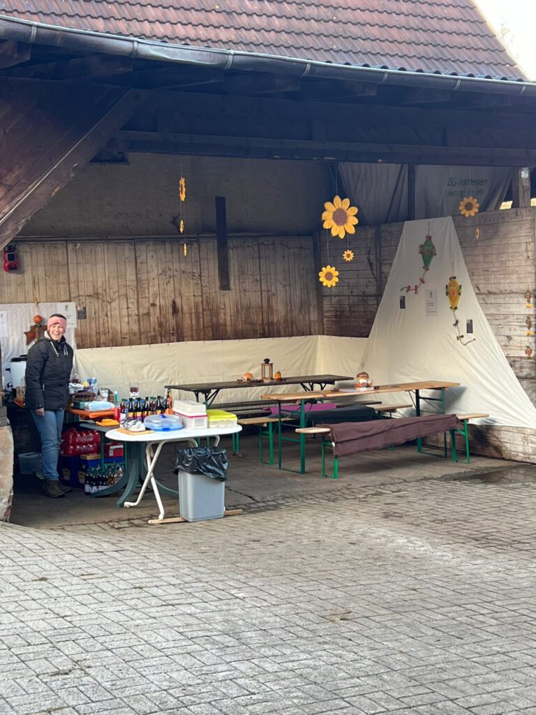Marktstand im Freien mit Tischen, Getränken und Sonnenblumendekorationen. Eine Person steht neben einer Kühlbox.