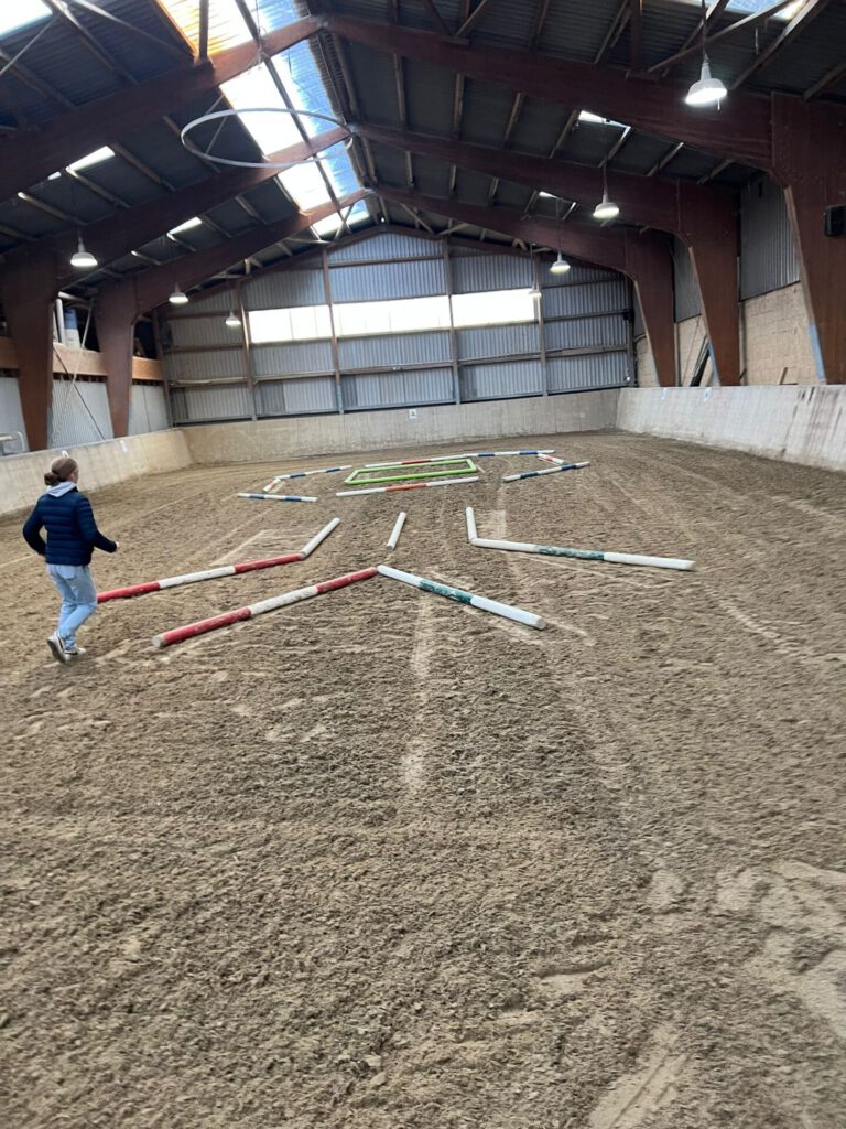 Indoor-Reithalle mit einer Person, die neben bunten Stangen auf dem Boden läuft.