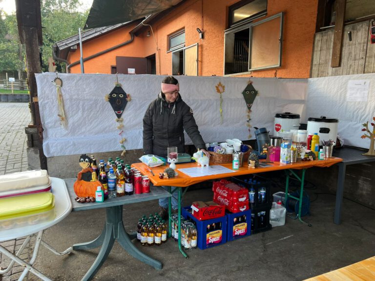 Eine Person steht hinter einem Tisch mit Getränken und Snacks im Freien mit herbstlicher Dekoration.