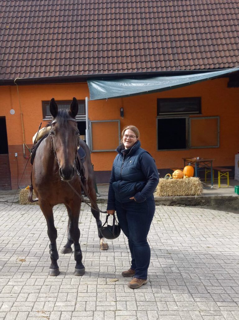 Eine Frau steht neben einem Pferd in einem Hof mit Heuballen und Kürbissen.